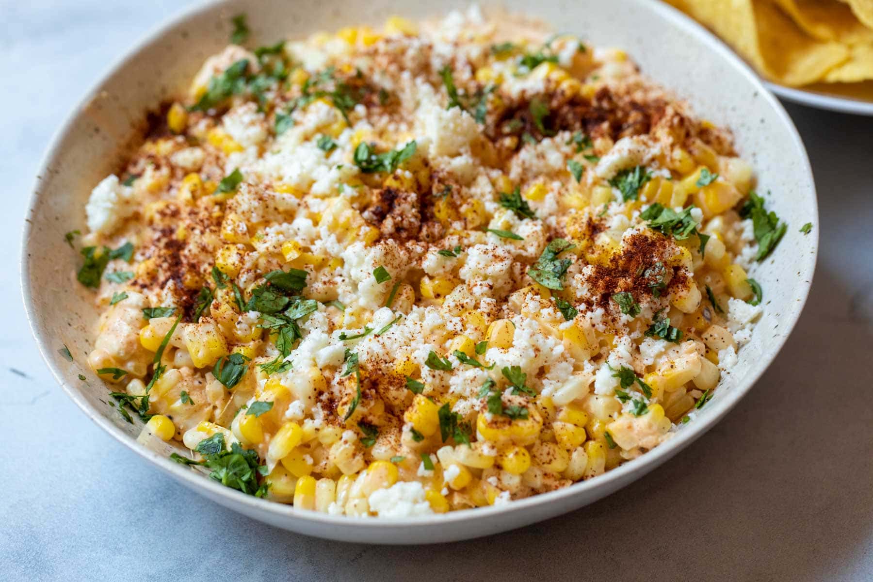 A rustic bowl of Mexican street corn dip topped with cotija cheese, chili powder, cilantro, and a lime wedge on a warm-toned kitchen counter with tortilla chips nearby