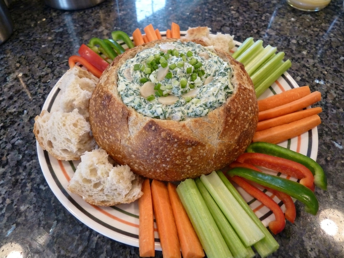 A rustic bread bowl filled with creamy Knorr vegetable dip, surrounded by torn bread pieces and fresh cut vegetables on a bright kitchen counter