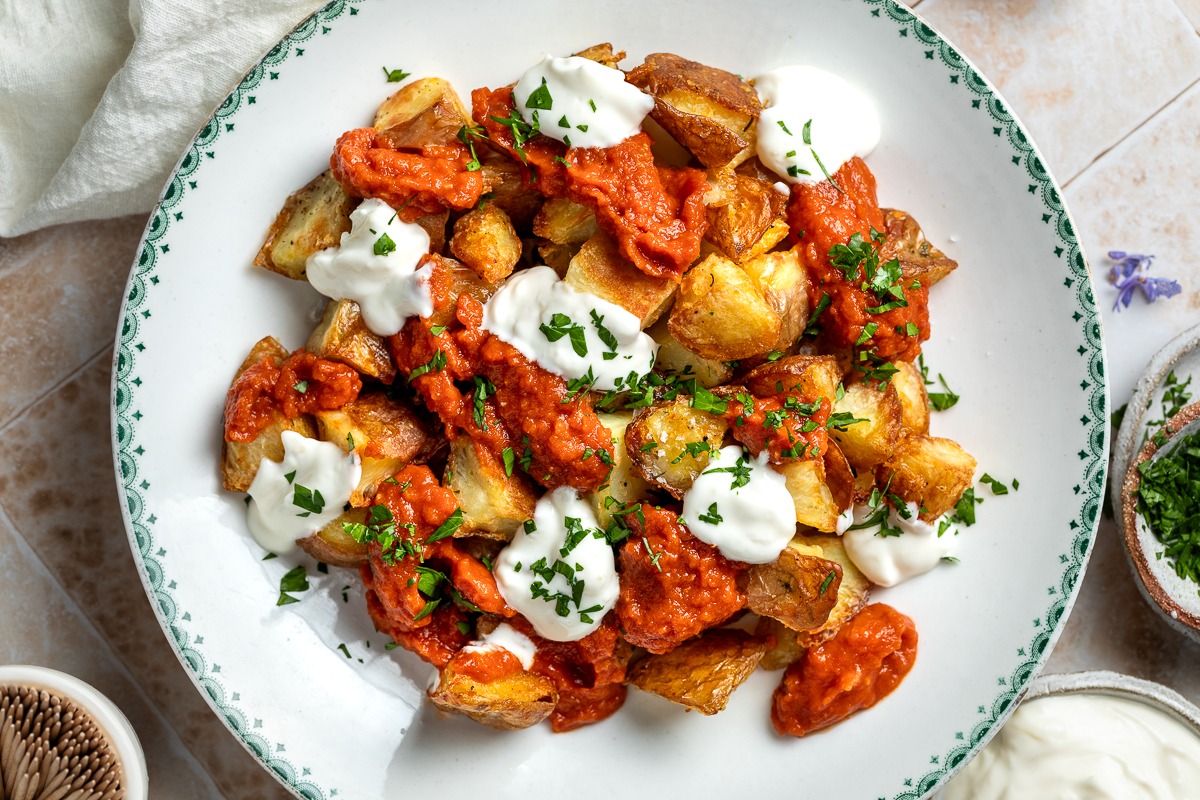 A rustic plate of patatas bravas with crispy golden potato cubes, smoky red bravas sauce spooned over the top, and a small bowl of garlic aioli on the side on a wooden table