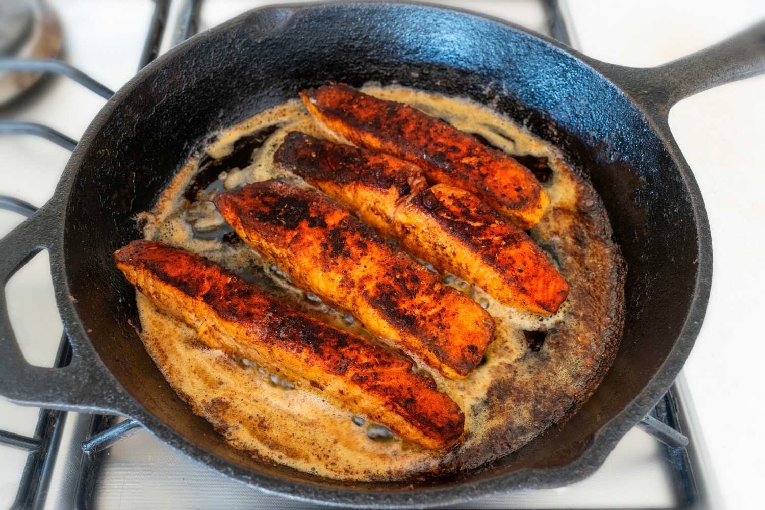 A salmon fillet cooking in a cast iron skillet with a dark blackened crust forming and a little butter bubbling around the edges