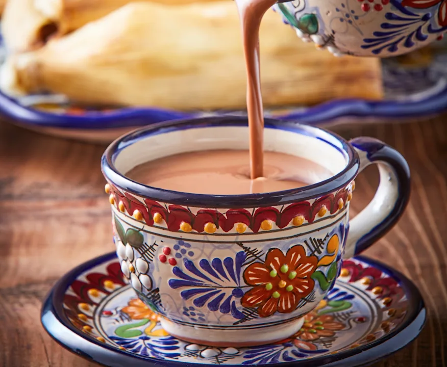 A saucepan of creamy atole being poured into a ceramic mug on a kitchen counter, with gentle steam rising