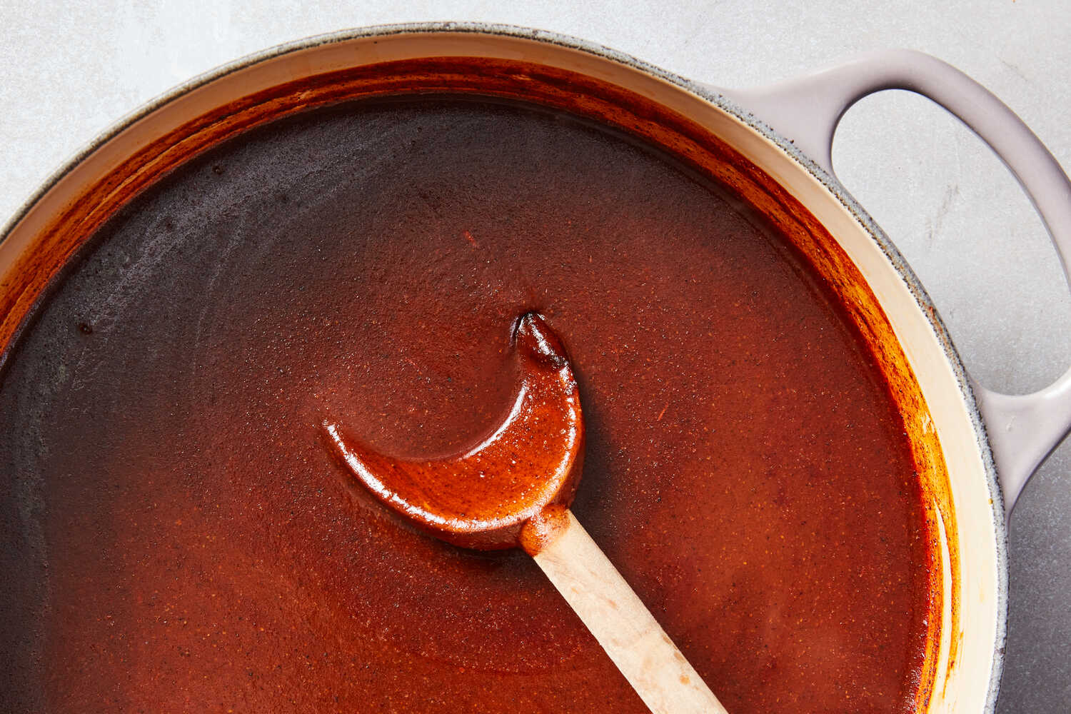 A saucepan of deep red enchilada sauce on a stovetop with a wooden spoon resting on the rim, warm kitchen lighting
