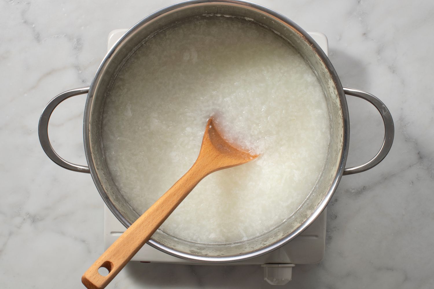 A saucepan on a home stove with leftover jook being reheated, a wooden spoon stirring while a small pitcher of broth sits nearby on the counter