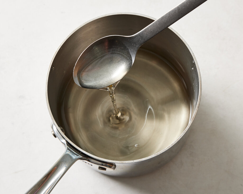 A saucepan on a stove with clear sugar syrup coming to a gentle boil as a wooden spoon stirs in a home kitchen