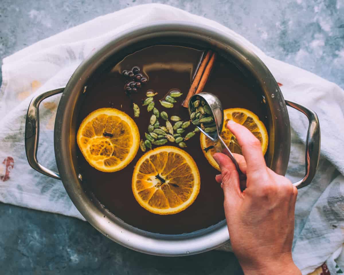 A saucepan on a stovetop with apple cider heating and whole spices plus orange peel floating on the surface