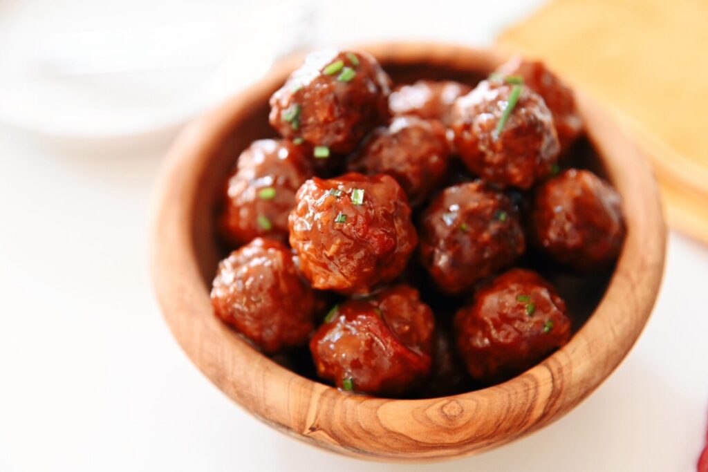 A saucepan on a stovetop with meatballs simmering in a glossy red-purple sauce while a wooden spoon stirs, warm kitchen lighting consistent with the other images
