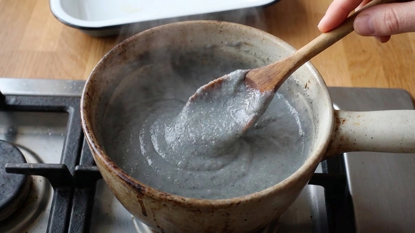 A saucepan on a stovetop with thickening cornmeal porridge being stirred with a wooden spoon as steam rises