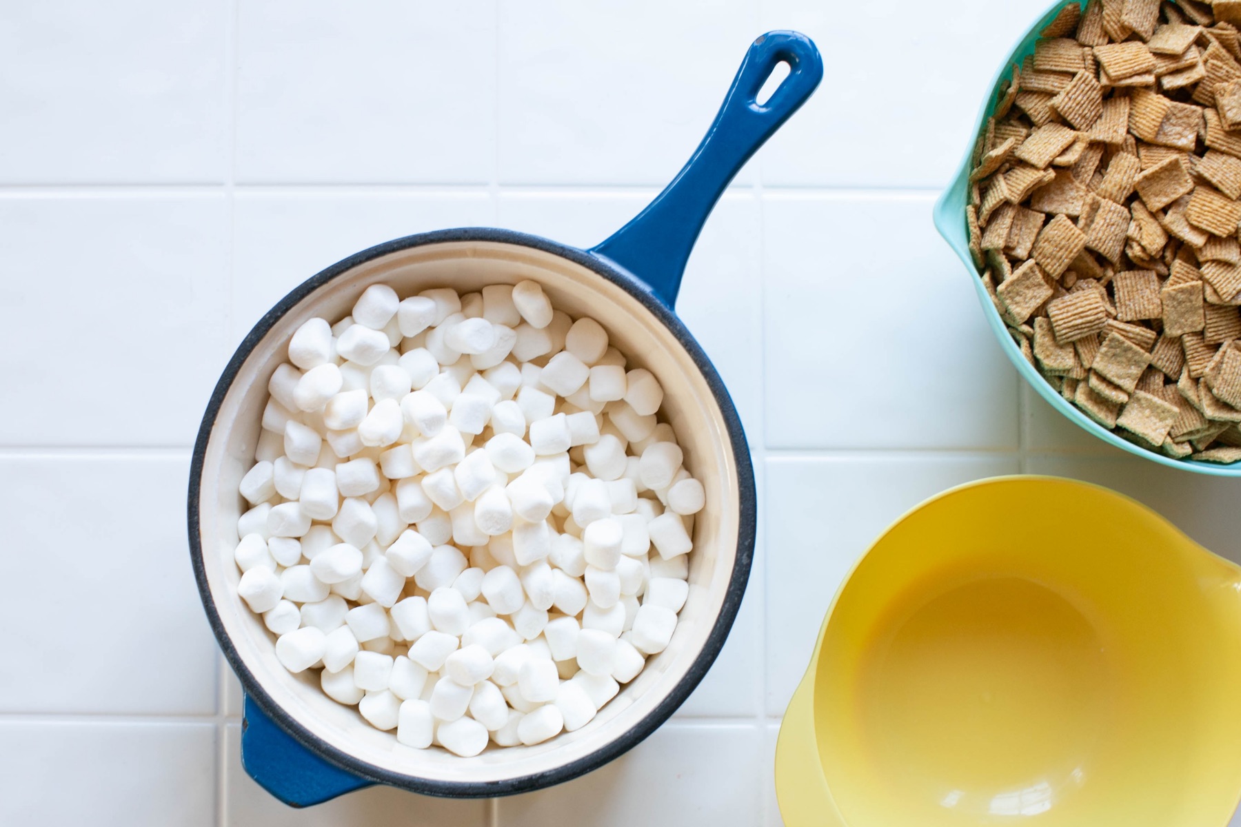 A saucepan surrounded by butter, mini marshmallows, vanilla extract, flaky salt, and crispy rice cereal on a clean kitchen counter