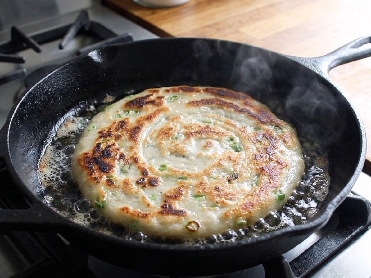 A scallion pancake frying in a cast iron skillet with bubbling oil and golden brown edges forming