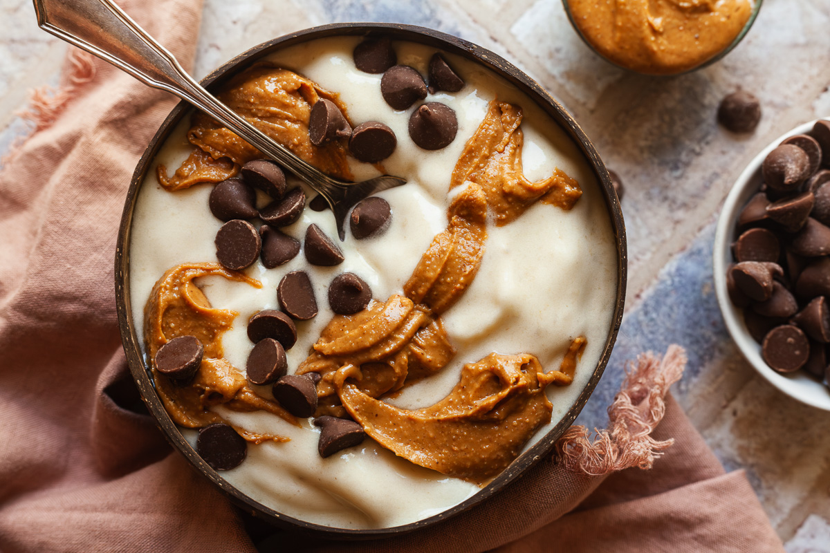 A scoop of creamy banana ice cream in a ceramic bowl on a light stone kitchen counter, topped with sliced banana and a sprinkle of cinnamon, soft natural window light