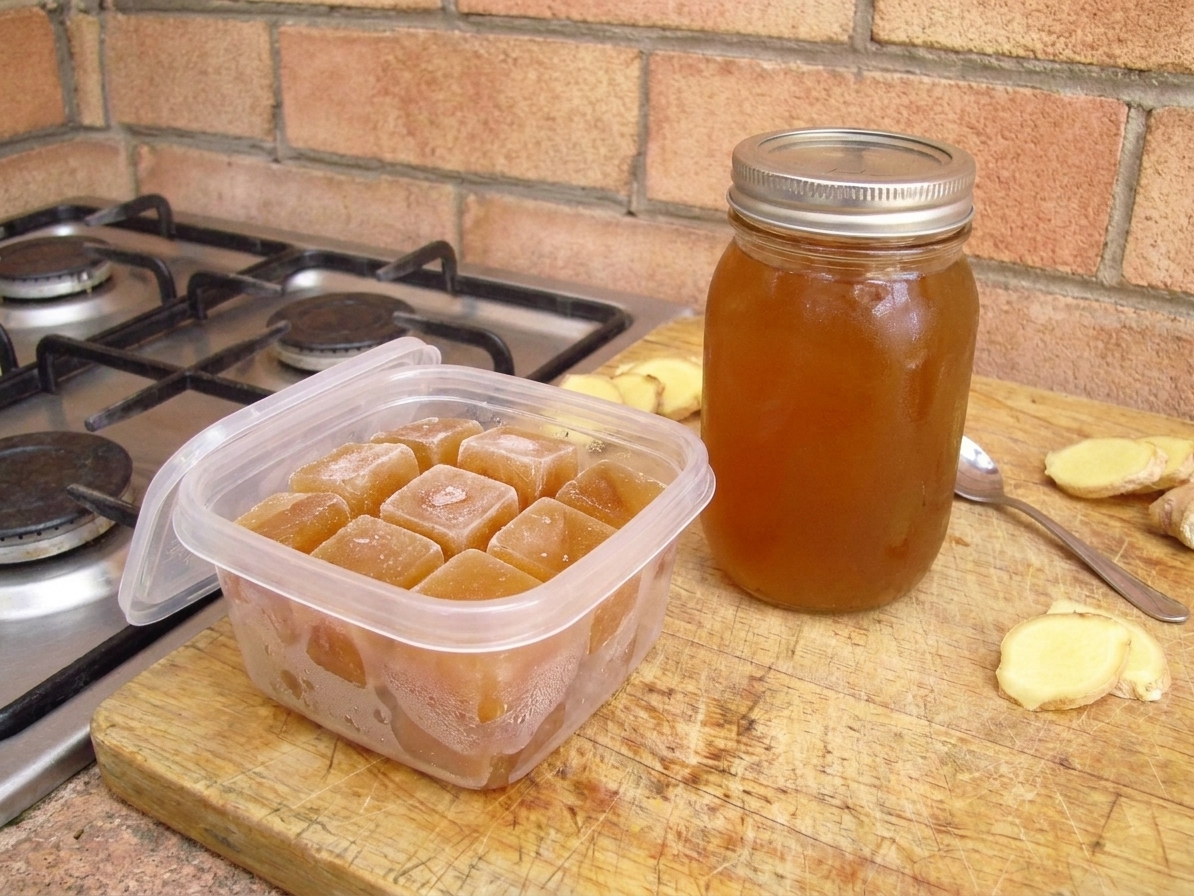 A sealed glass bottle of ginger syrup next to a small jar of ginger syrup cubes in a freezer-safe container on a kitchen counter