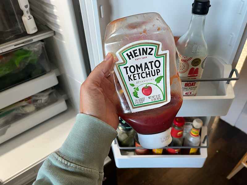 A sealed glass jar of homemade ketchup sitting on a refrigerator shelf next to common condiments