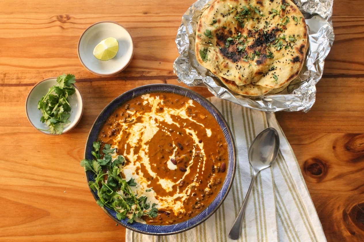 A serving bowl of dal makhni on a dinner table with naan, a spoon, and small bowls of cilantro and lemon wedges in warm evening light