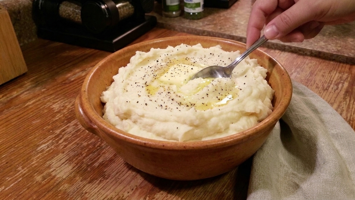 A serving bowl of whipped potatoes with a pat of butter melting into the surface while a spoon makes soft swirls