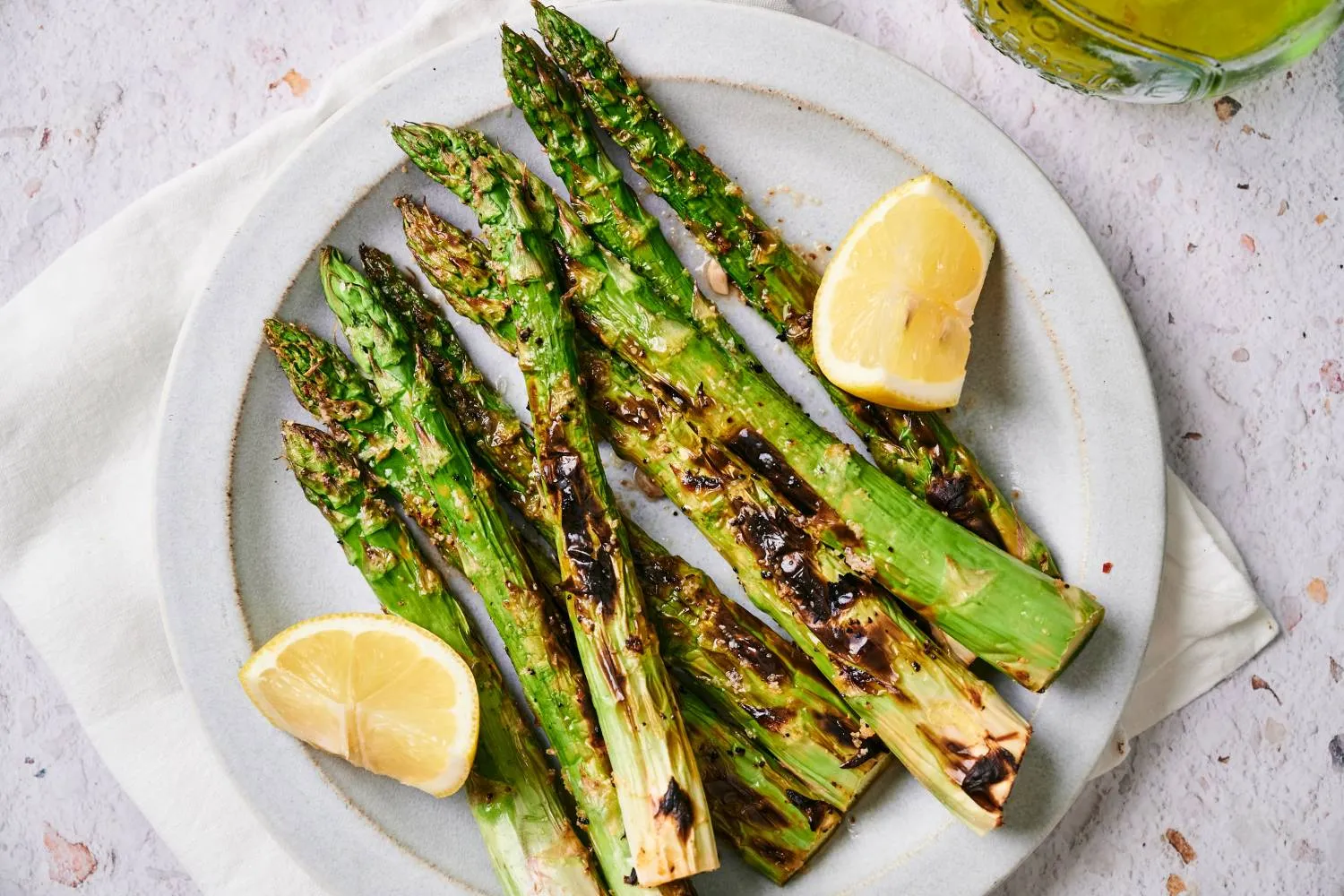 A serving platter of grilled asparagus with light char marks, lemon wedges, and flaky salt on a wooden table in a bright kitchen