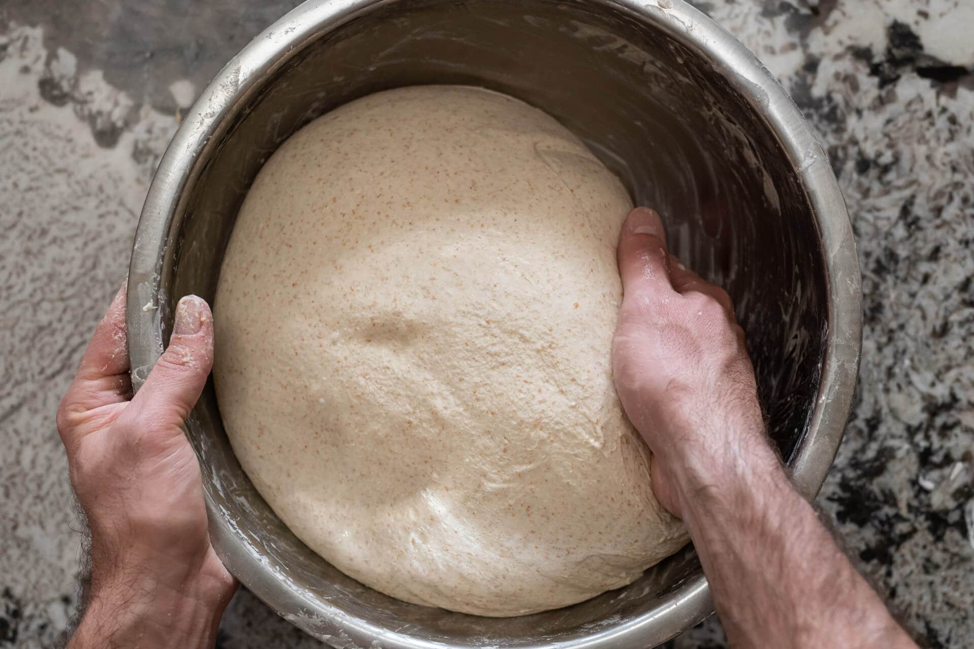 A shaggy whole wheat bread dough being mixed in a large bowl with a wooden spoon on a kitchen counter