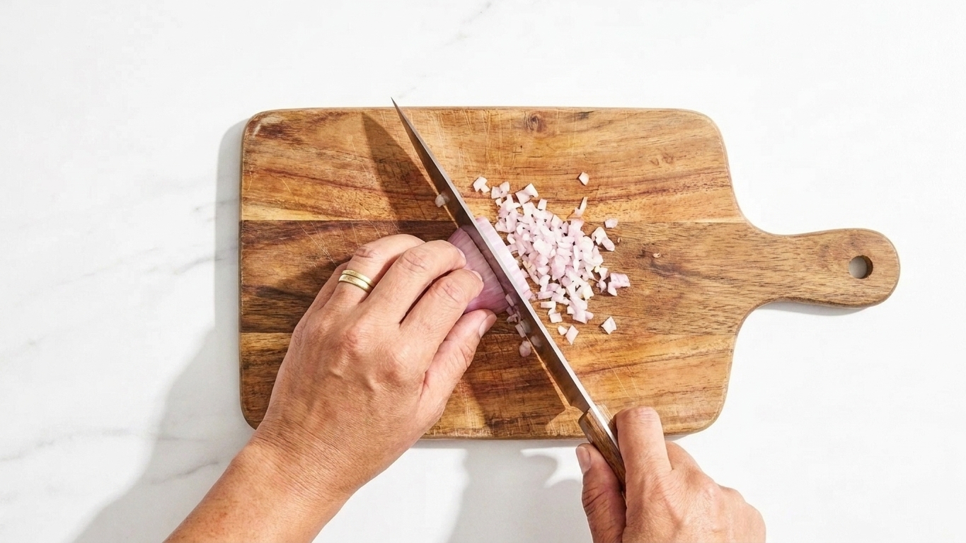 A shallot being finely minced on a wooden cutting board with a chef's knife on a bright kitchen counter