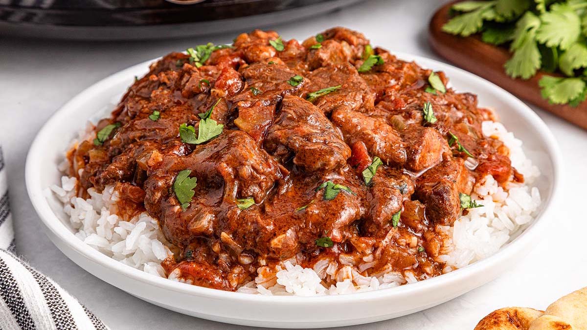 A shallow bowl of beef curry with tender chunks of beef in a rich reddish-brown sauce, served over fluffy white rice with cilantro on top, on a warmly lit kitchen table