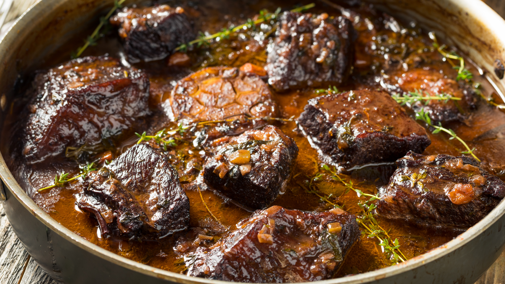 A shallow bowl of braised boneless beef short ribs covered in glossy red wine gravy, served with mashed potatoes on a rustic table with a linen napkin