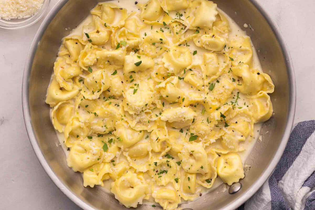 A shallow bowl of cheese tortellini coated in creamy Parmesan sauce, finished with black pepper and chopped parsley on a rustic wooden table