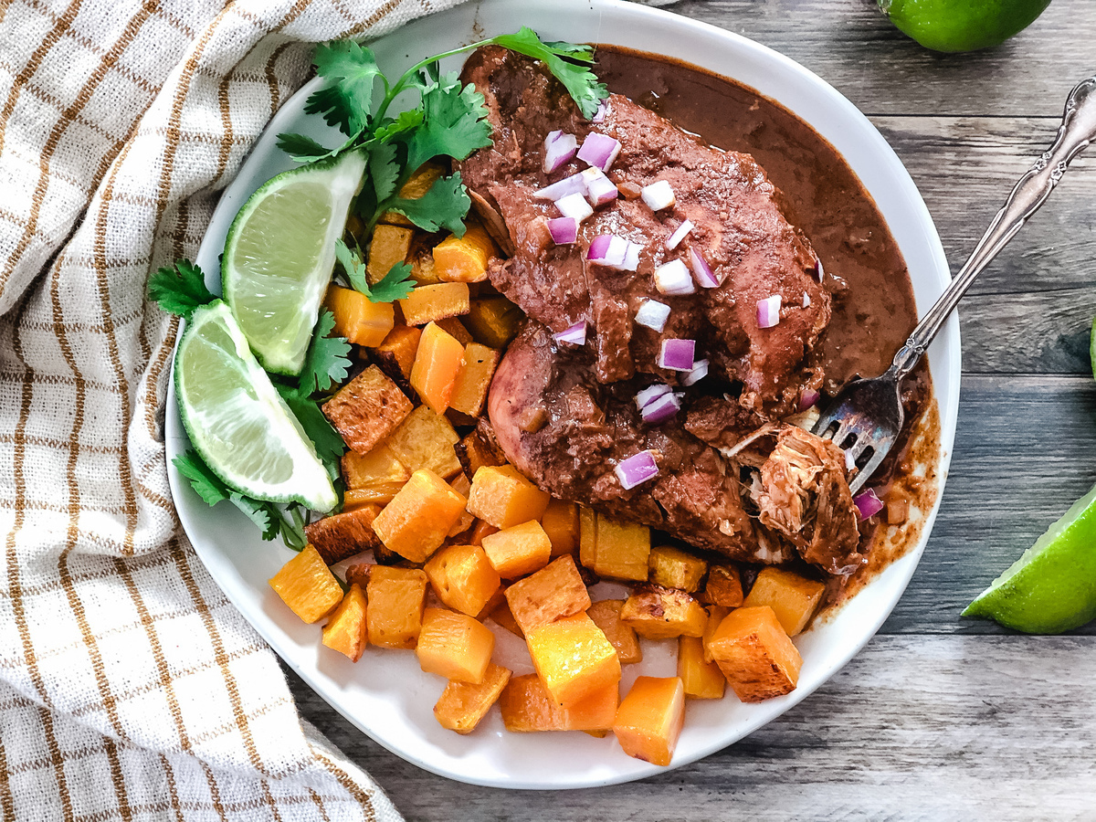 A shallow bowl of chicken mole with glossy dark sauce, sprinkled with toasted sesame seeds and chopped cilantro, served with warm tortillas on a wooden table in soft natural light