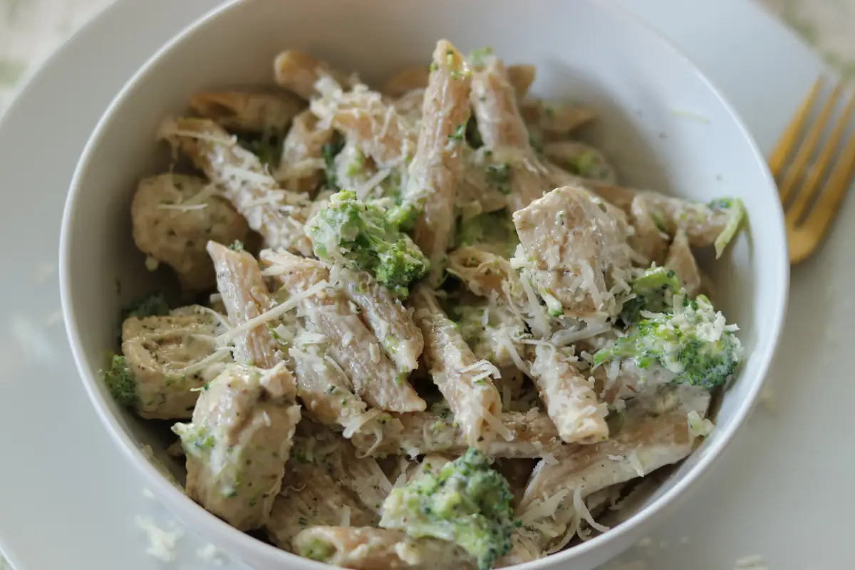 A shallow bowl of creamy chicken broccoli alfredo with Parmesan on top, set on a warm wood table with a fork and linen napkin, soft natural kitchen light