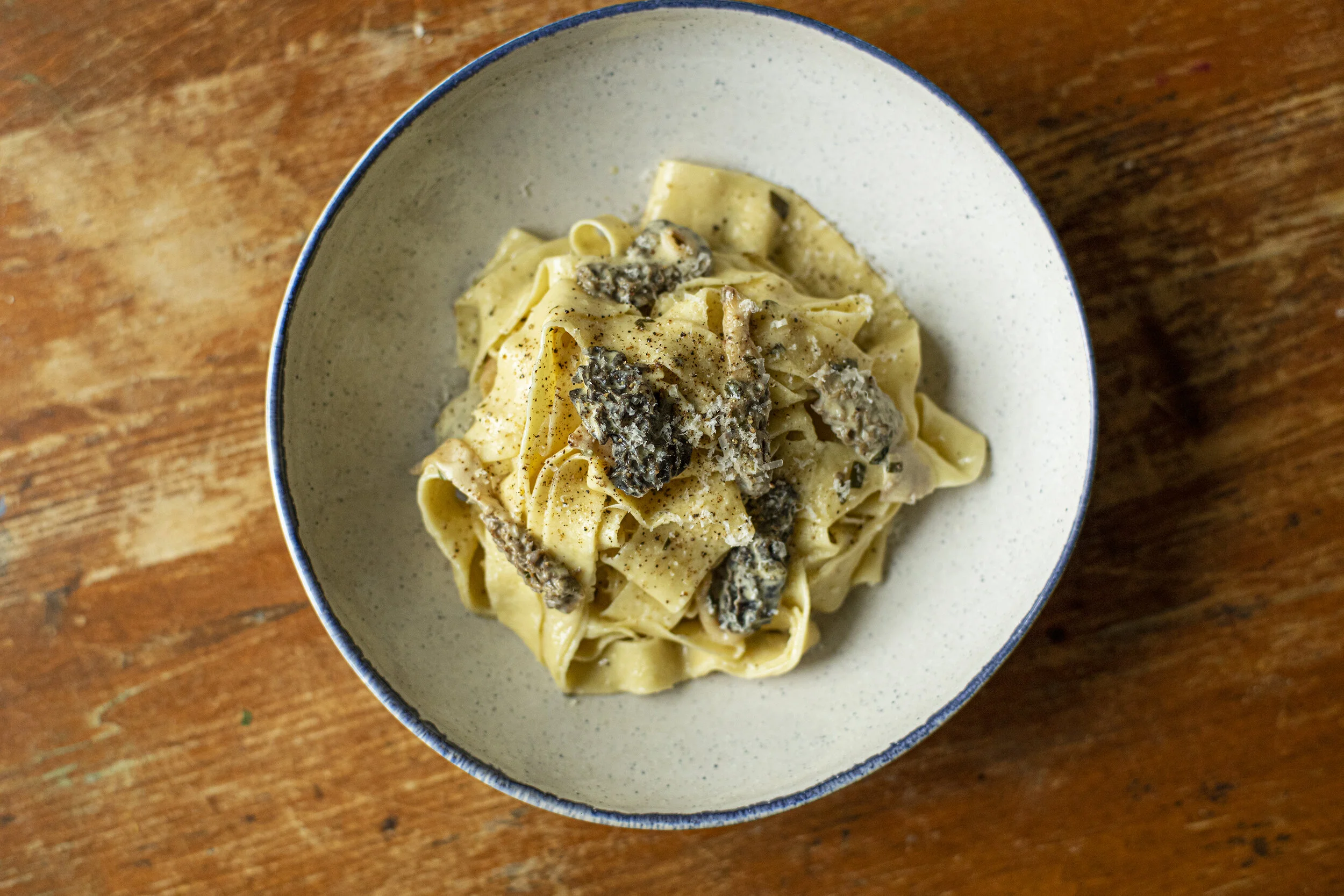 A shallow bowl of creamy morel mushroom pasta with visible sliced morels, grated Parmesan, black pepper, and lemon zest on a rustic wooden table with a fork beside it