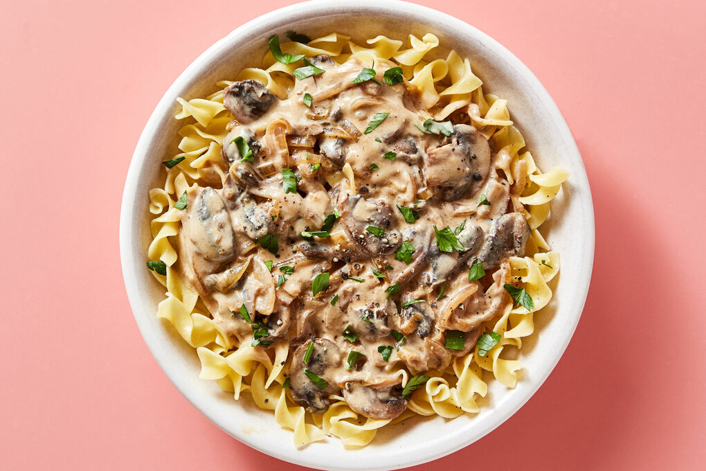 A shallow bowl of creamy mushroom stroganoff over egg noodles, garnished with chopped parsley on a warm wooden table with soft natural light