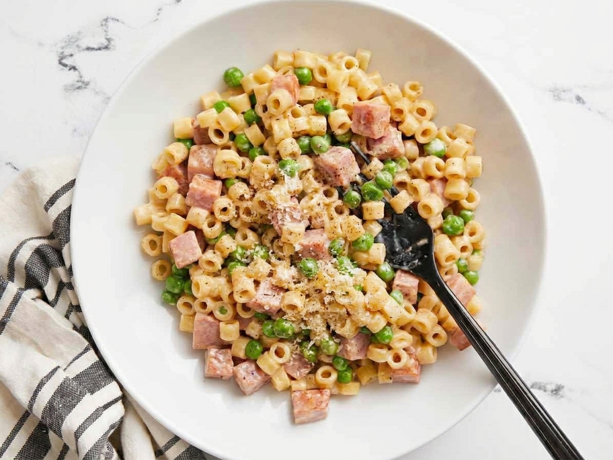 A shallow bowl of creamy pasta with diced ham and green peas, finished with grated Parmesan and black pepper on a warm kitchen table