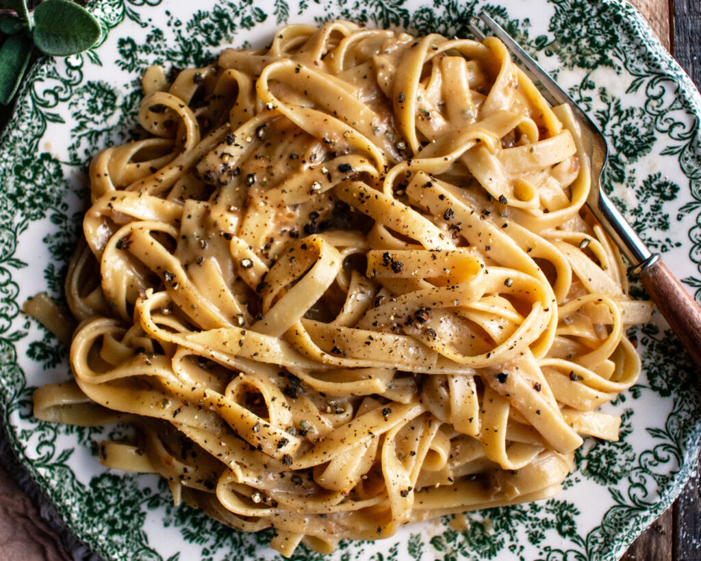 A shallow bowl of fettuccine coated in glossy Alfredo sauce, topped with freshly grated Parmesan and black pepper on a warm wood table
