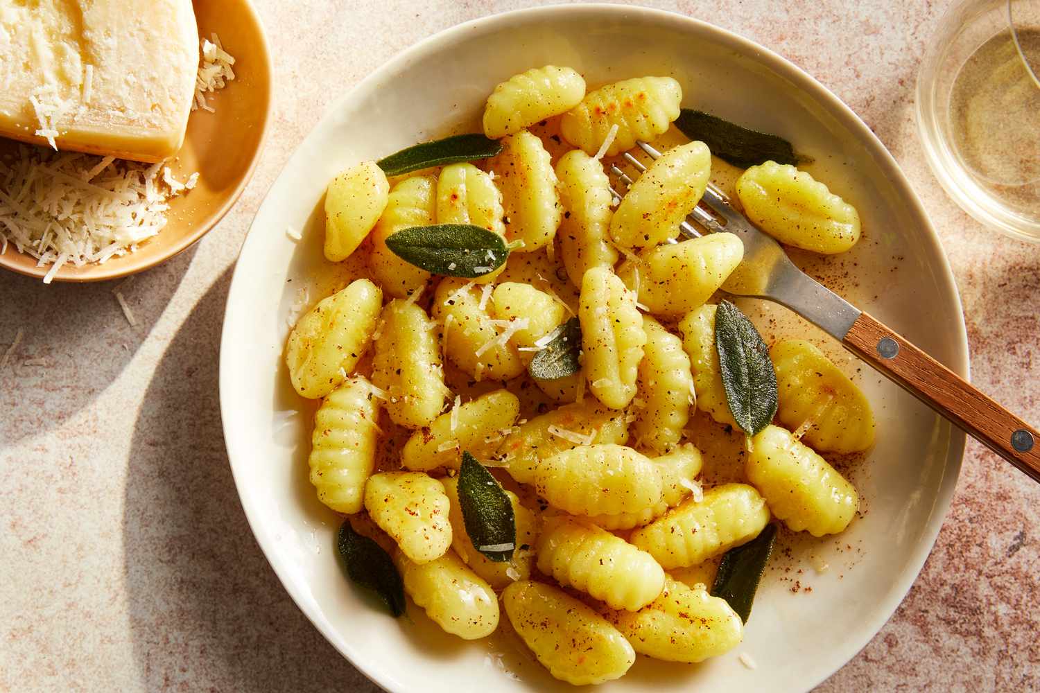 A shallow bowl of homemade potato gnocchi coated in browned butter with crispy sage leaves and a dusting of Parmesan on a rustic wooden table