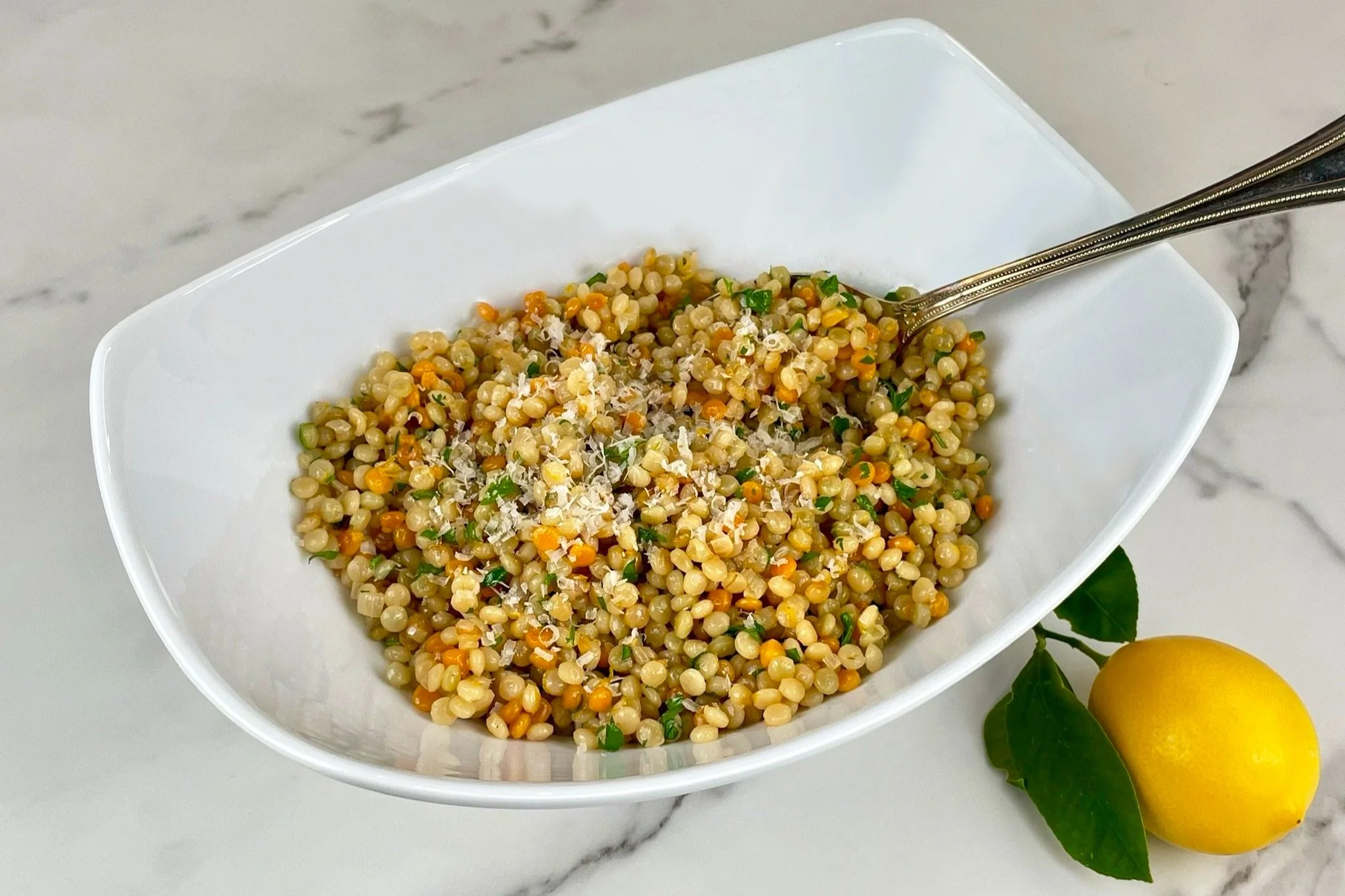 A shallow bowl of lemony pearl couscous with chopped parsley and toasted pine nuts on a light wooden table, warm natural kitchen lighting
