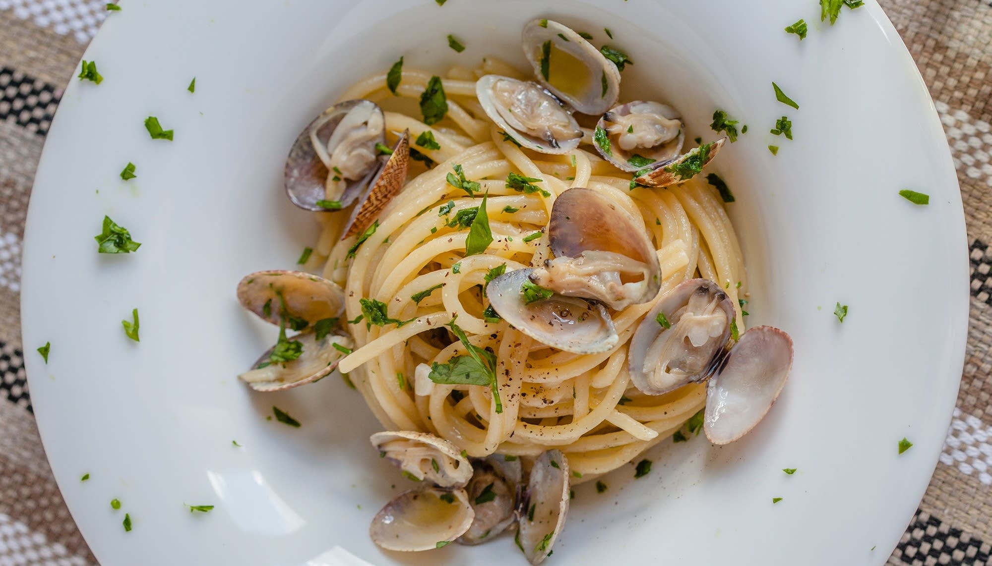 A shallow bowl of linguine with white clam sauce, chopped parsley, lemon zest, and a light sheen of olive oil on a wooden table with a fork beside it