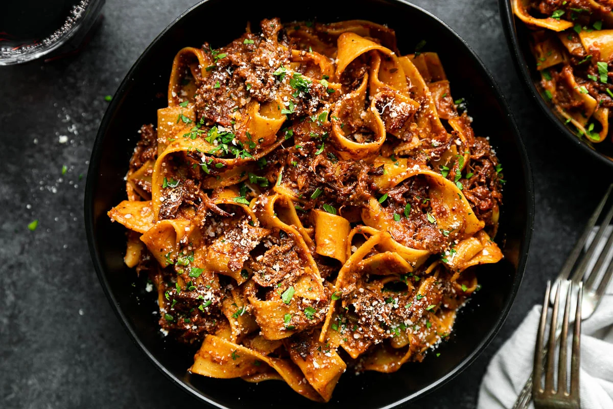 A shallow bowl of pappardelle tossed in short rib ragu, topped with grated Parmesan and chopped parsley on a warm-toned dinner table