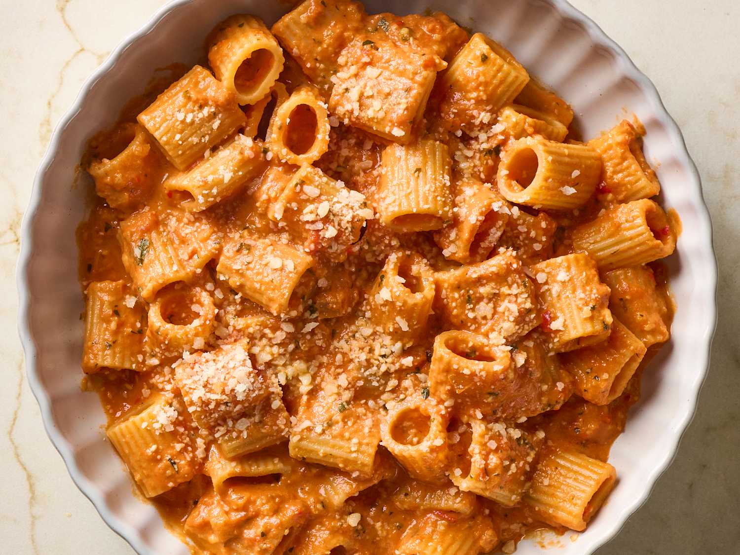A shallow bowl of rigatoni coated in creamy spicy tomato sauce, topped with grated parmesan and basil on a warm-toned kitchen table