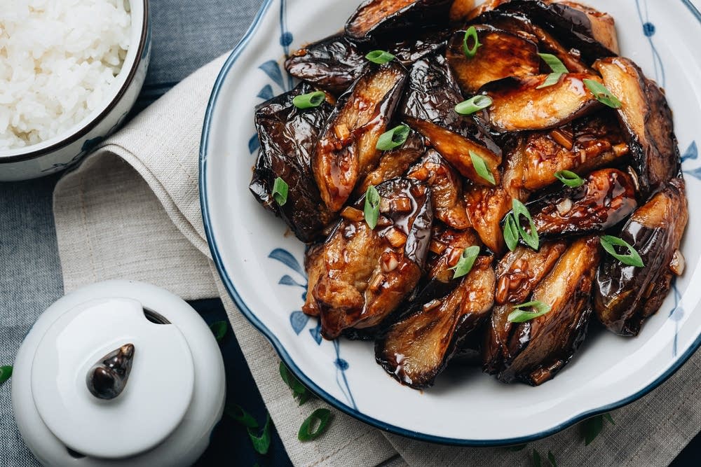 A shallow bowl of spicy garlic Chinese eggplant in a glossy reddish-brown sauce, topped with sliced scallions and sesame seeds, set on a warm, neutral-toned dinner table with chopsticks