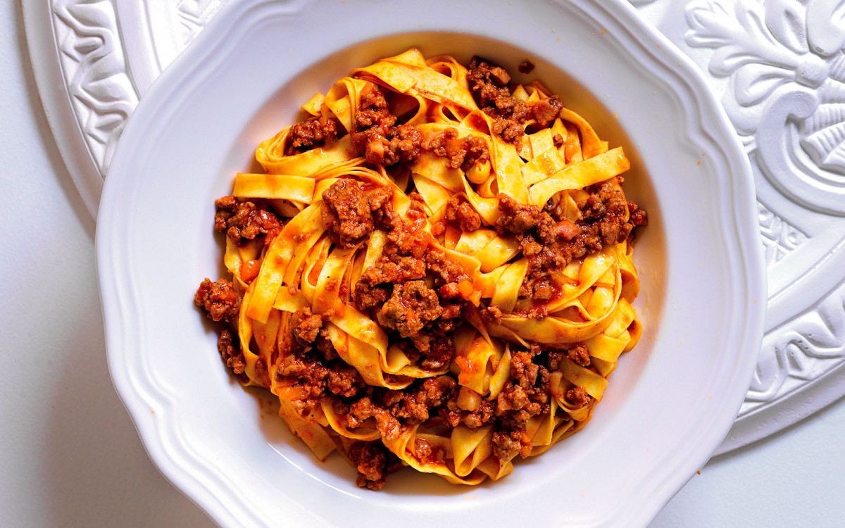 A shallow bowl of tagliatelle topped with bolognese sauce and finely grated Parmesan on a wooden table with a simple linen napkin