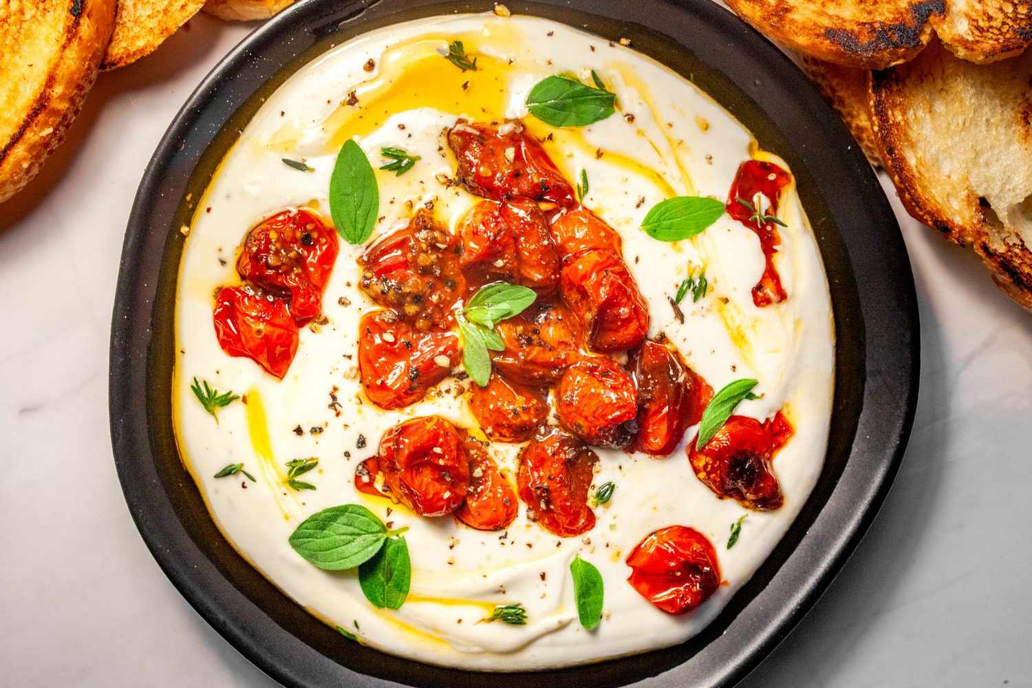 A shallow bowl of whipped feta swirled with olive oil and topped with cracked black pepper and fresh herbs on a light stone kitchen counter