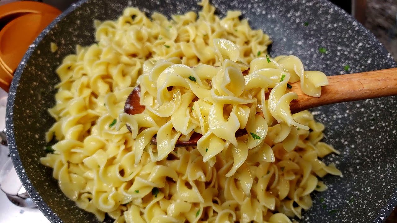 A shallow skillet filled with glossy buttered egg noodles sprinkled with black pepper and parsley on a warm kitchen counter