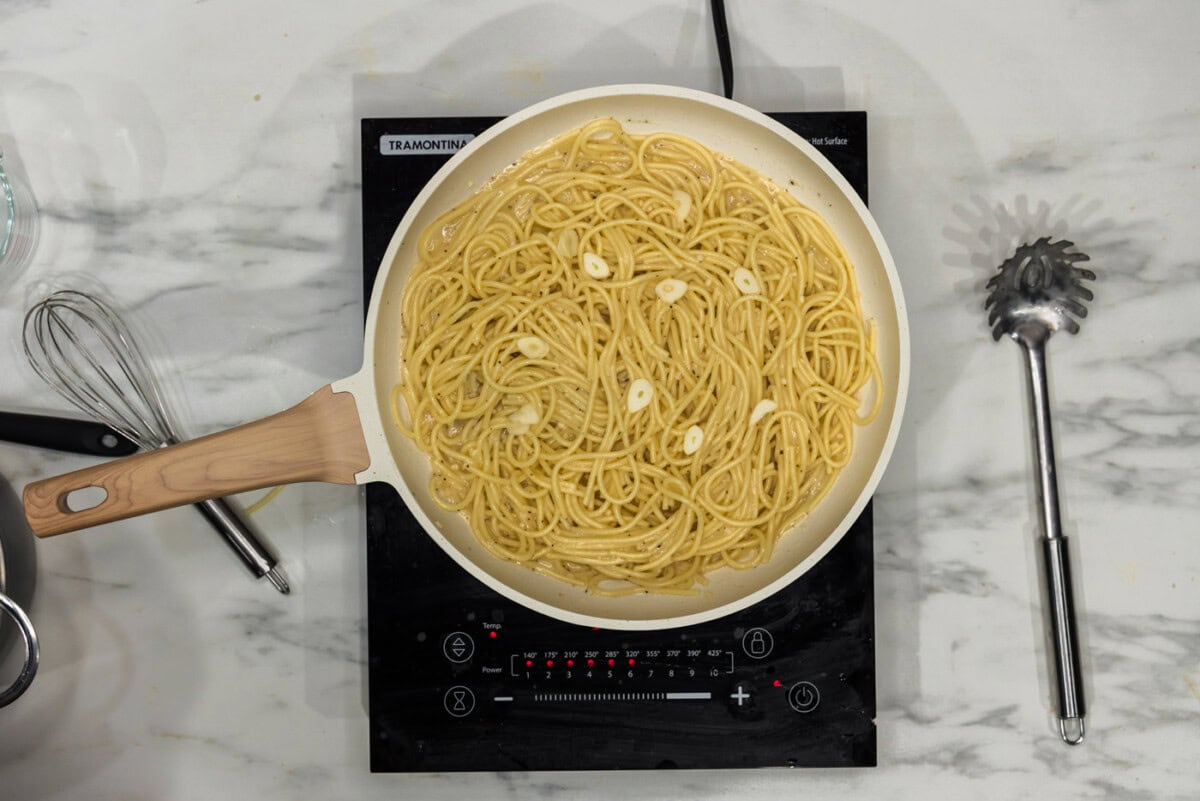 A shallow skillet filled with glossy garlic butter pasta, speckled with black pepper and parmesan, with a lemon wedge on the side on a warm kitchen counter