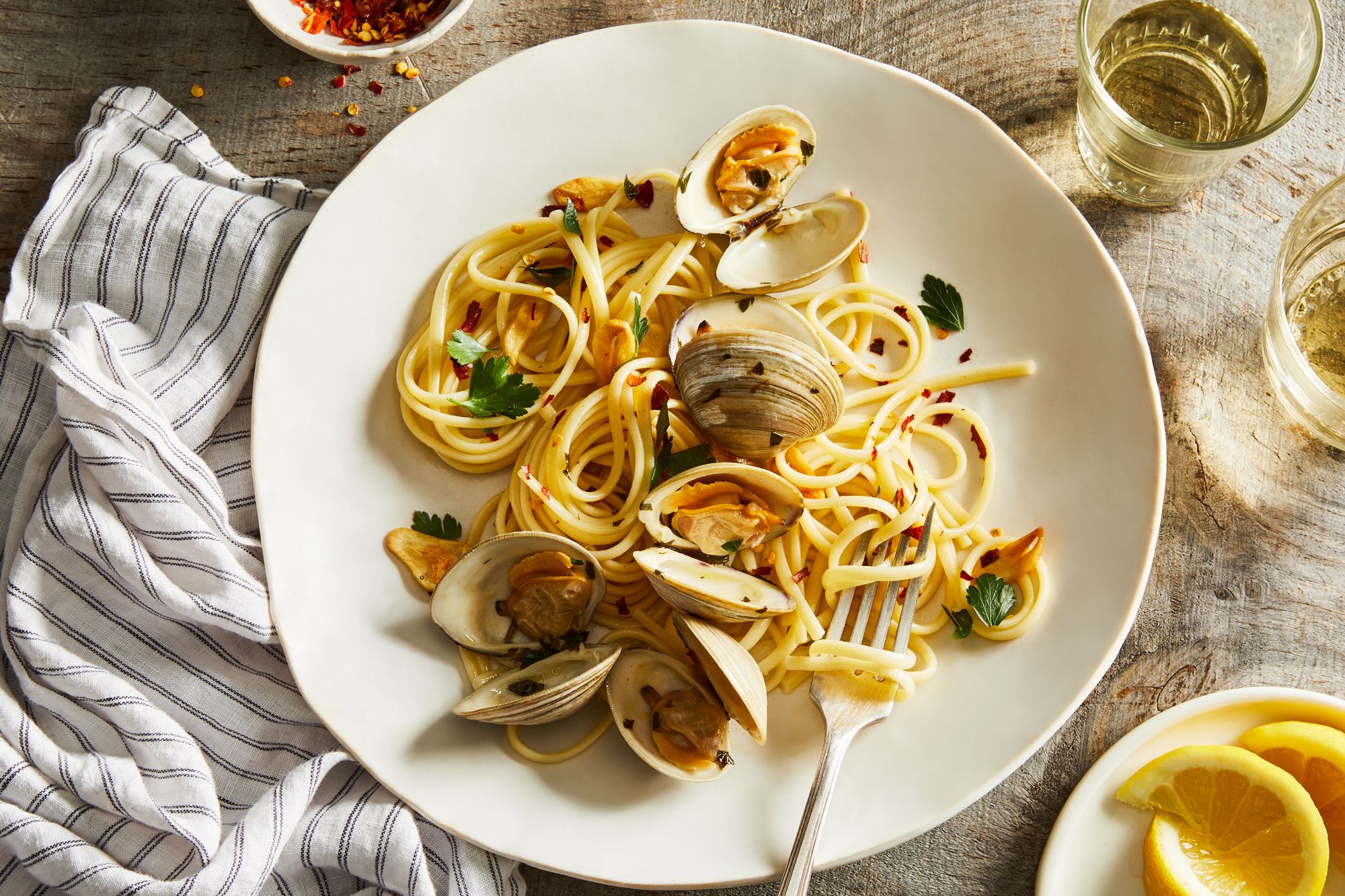 A shallow white bowl of clam linguine with chopped parsley, lemon zest, and clam shells on a light wood table with a linen napkin