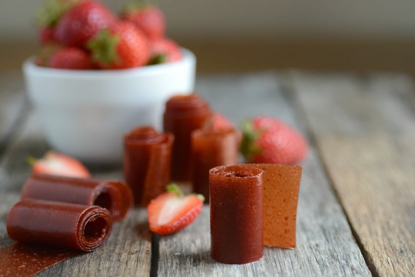 A sheet of glossy strawberry fruit leather peeled back on parchment on a kitchen counter, with a few cut roll-ups on the side