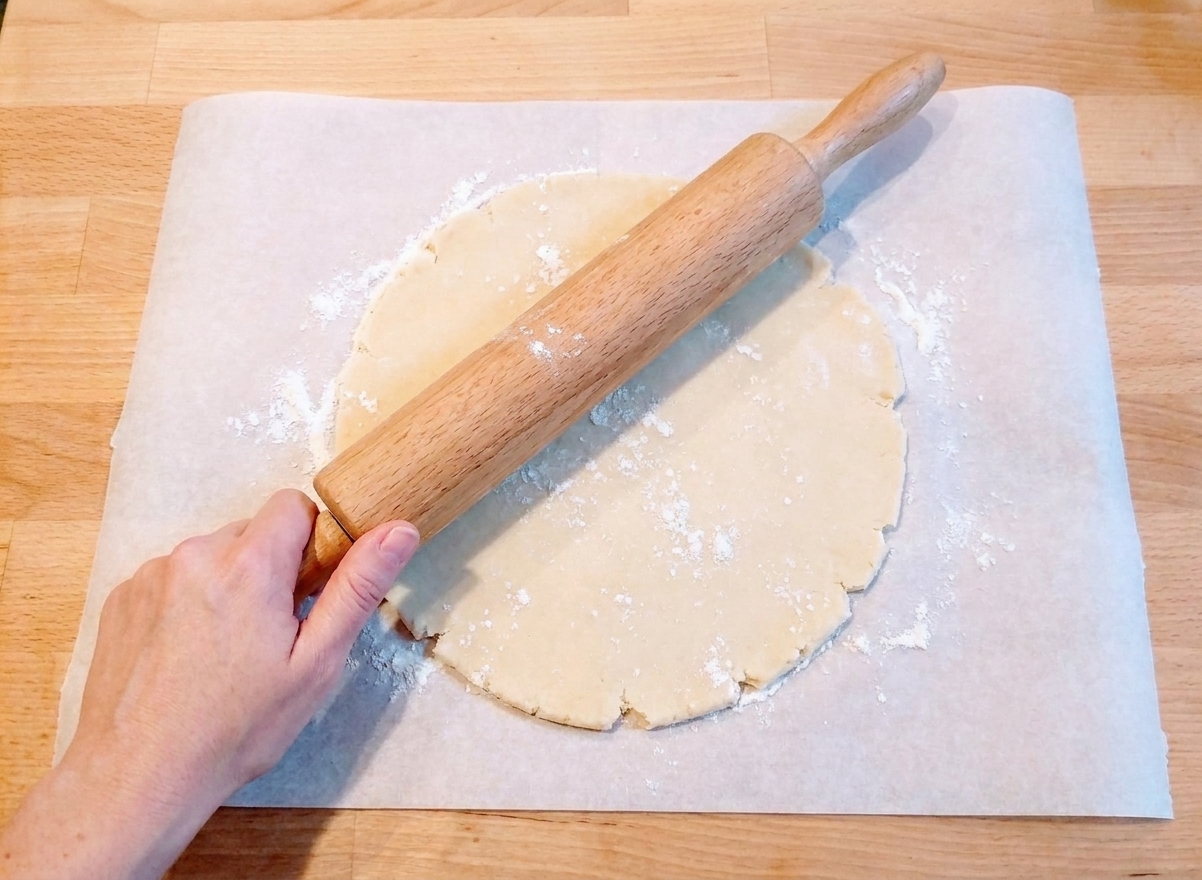 A sheet of parchment paper on a kitchen counter with pie dough being rolled out using a wooden rolling pin