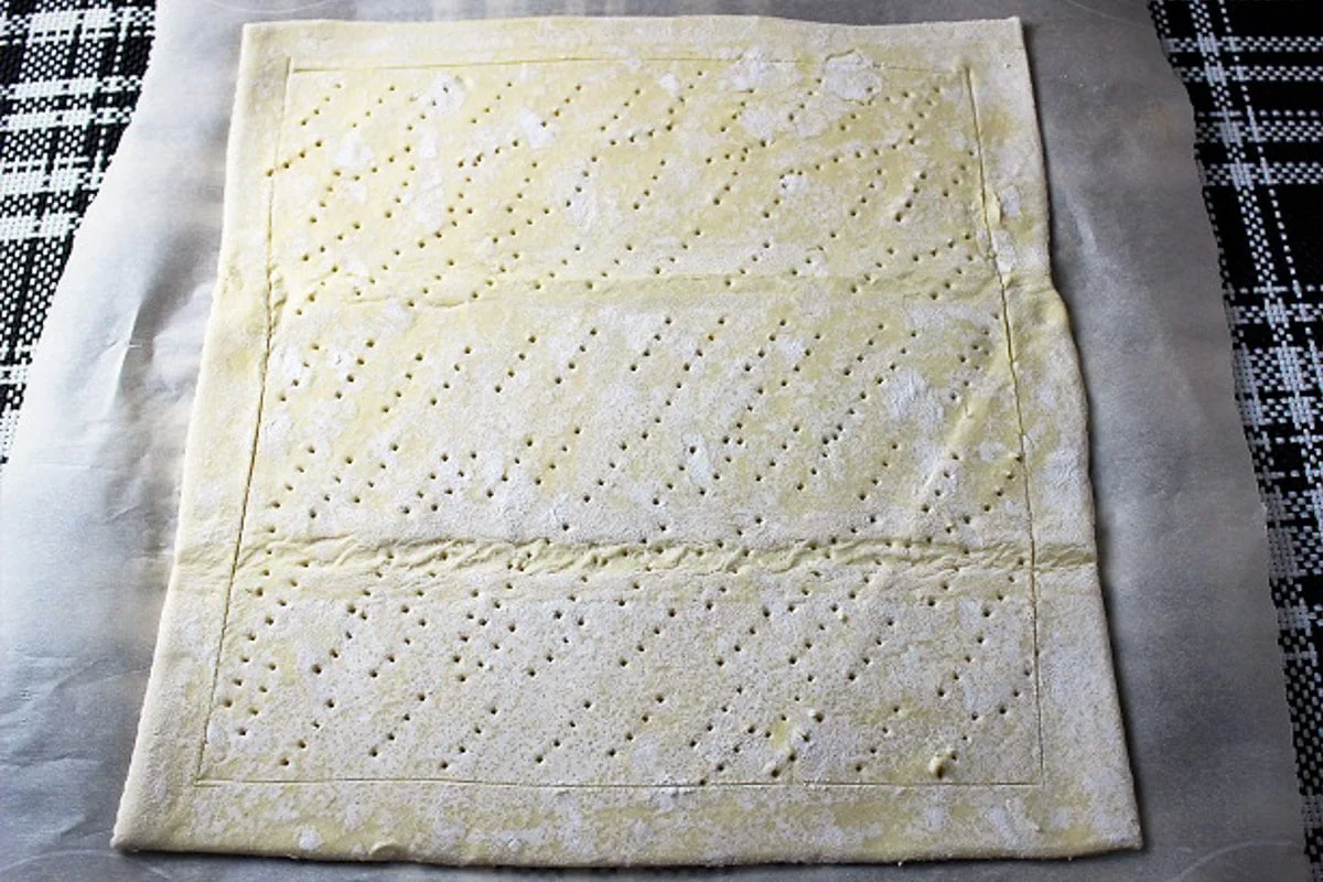 A sheet of puff pastry on parchment paper being scored with a shallow border using a small knife