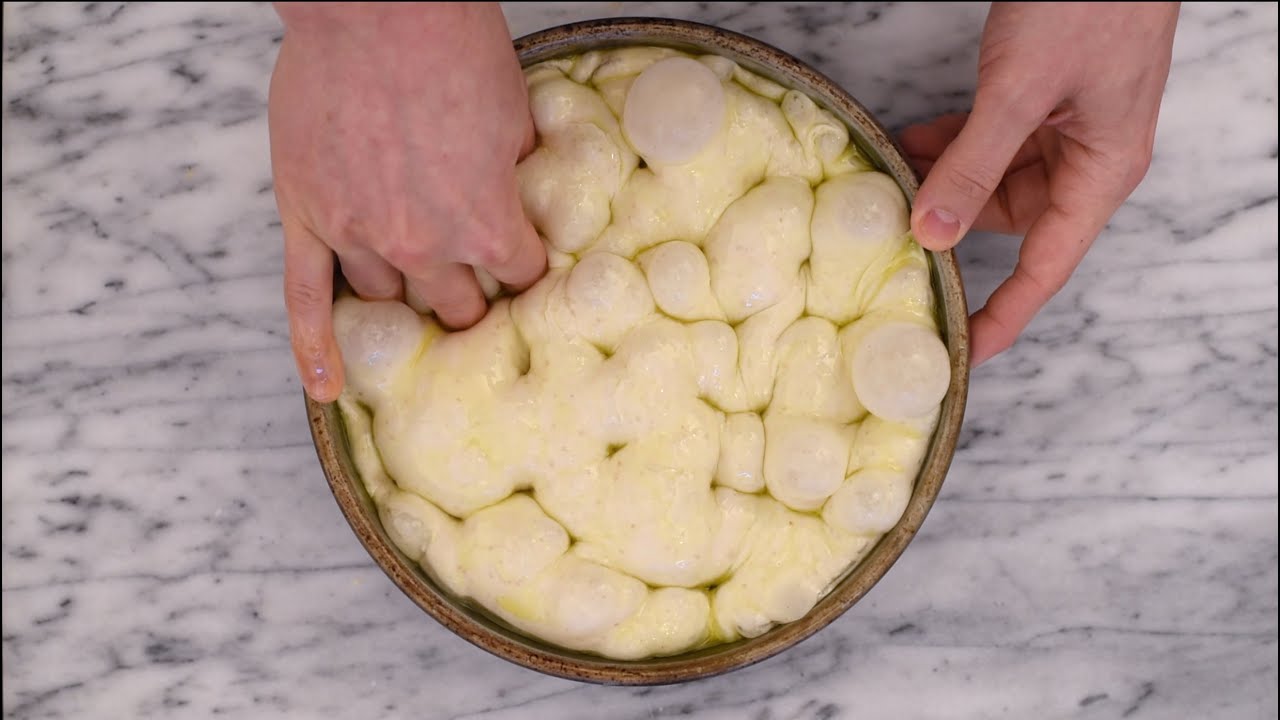A sheet pan filled with risen focaccia dough being dimpled with fingertips, with olive oil glistening in the indentations