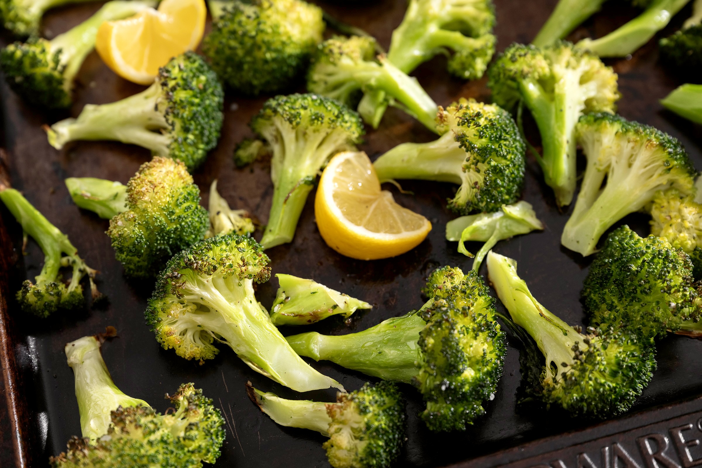 A sheet pan of crispy roasted broccoli florets with browned edges and lemon wedges on the side