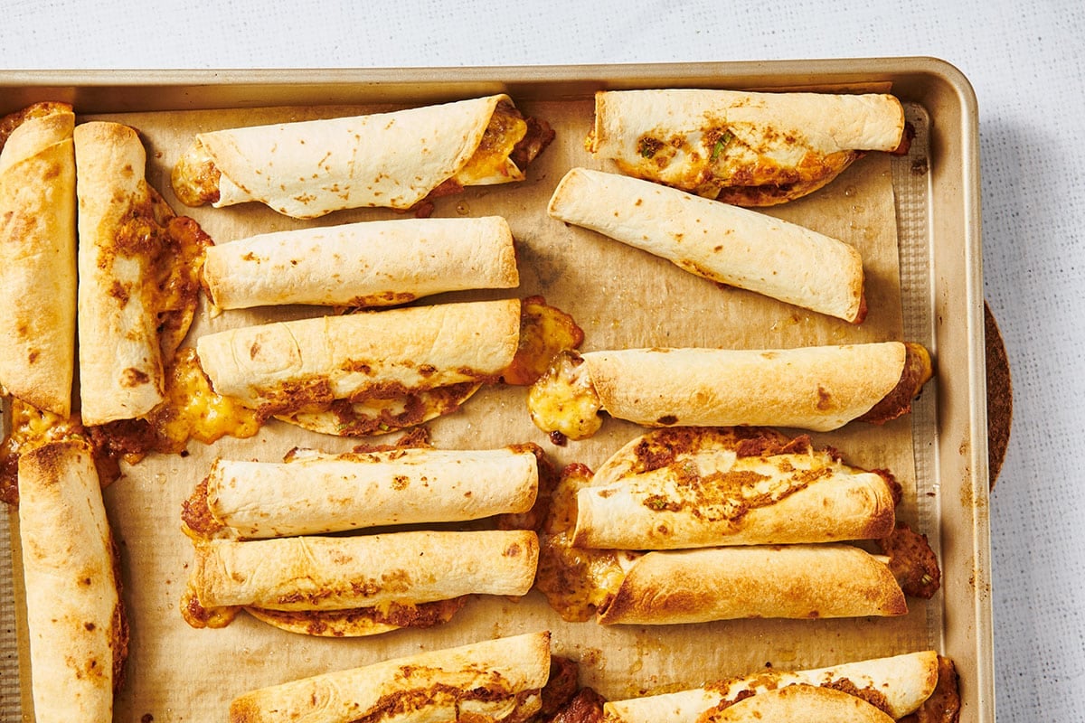 A sheet pan of flautas arranged in a single layer, lightly oiled and ready to go into the oven on a home kitchen counter