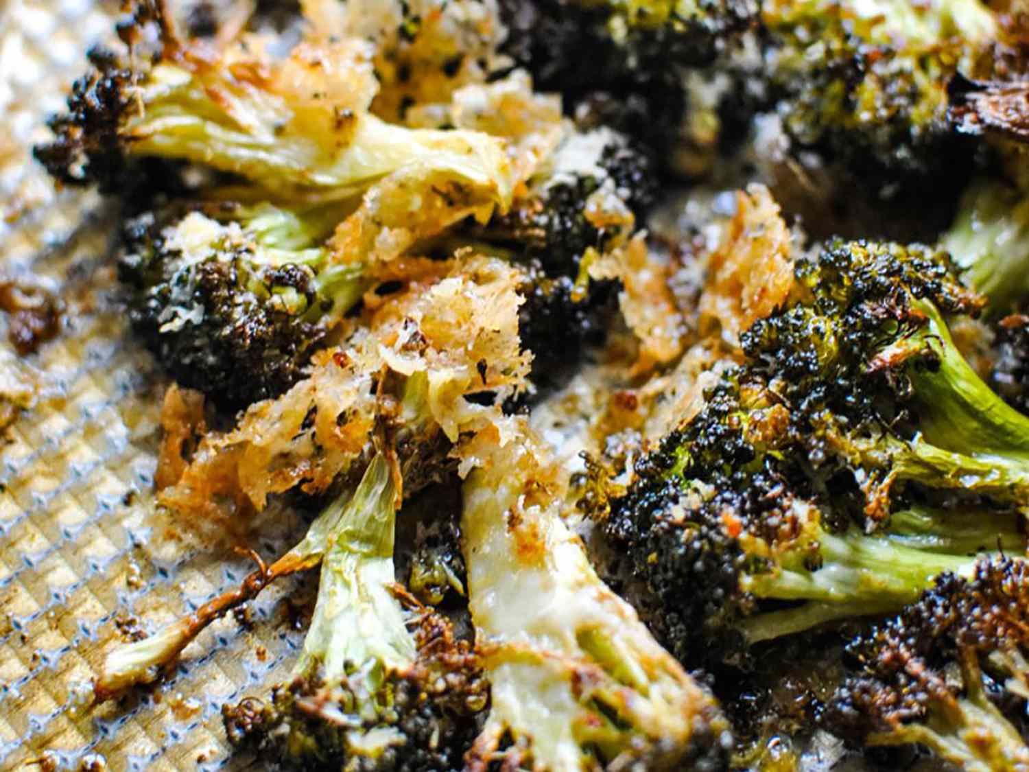 A sheet pan of oven roasted broccoli with deeply browned crisp edges, scattered parmesan, and lemon wedges on a warm kitchen counter