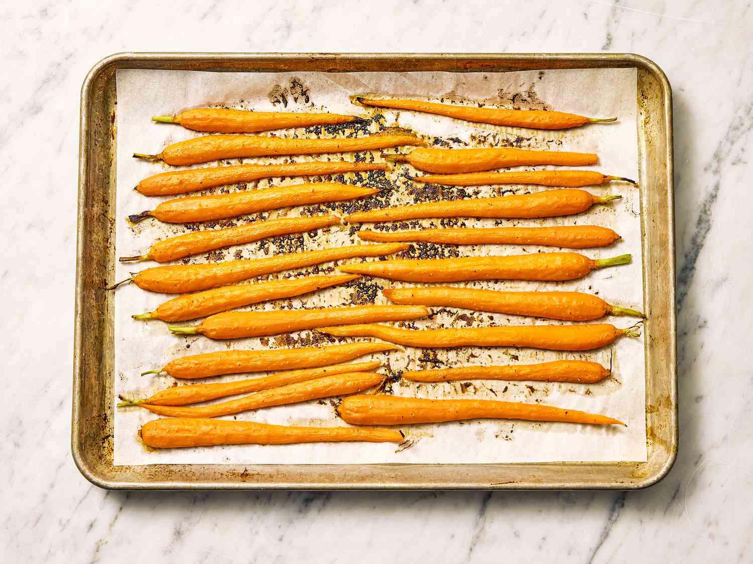 A sheet pan of oven roasted carrots with caramelized edges, glistening with a honey butter glaze, on a warm-toned kitchen counter