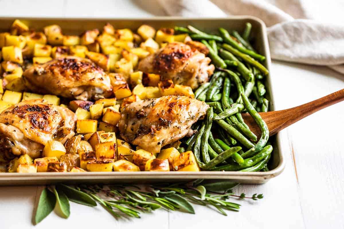 A sheet pan of roasted chicken thighs and golden baby potatoes with browned edges, scattered with herbs and lemon slices on a warm kitchen counter